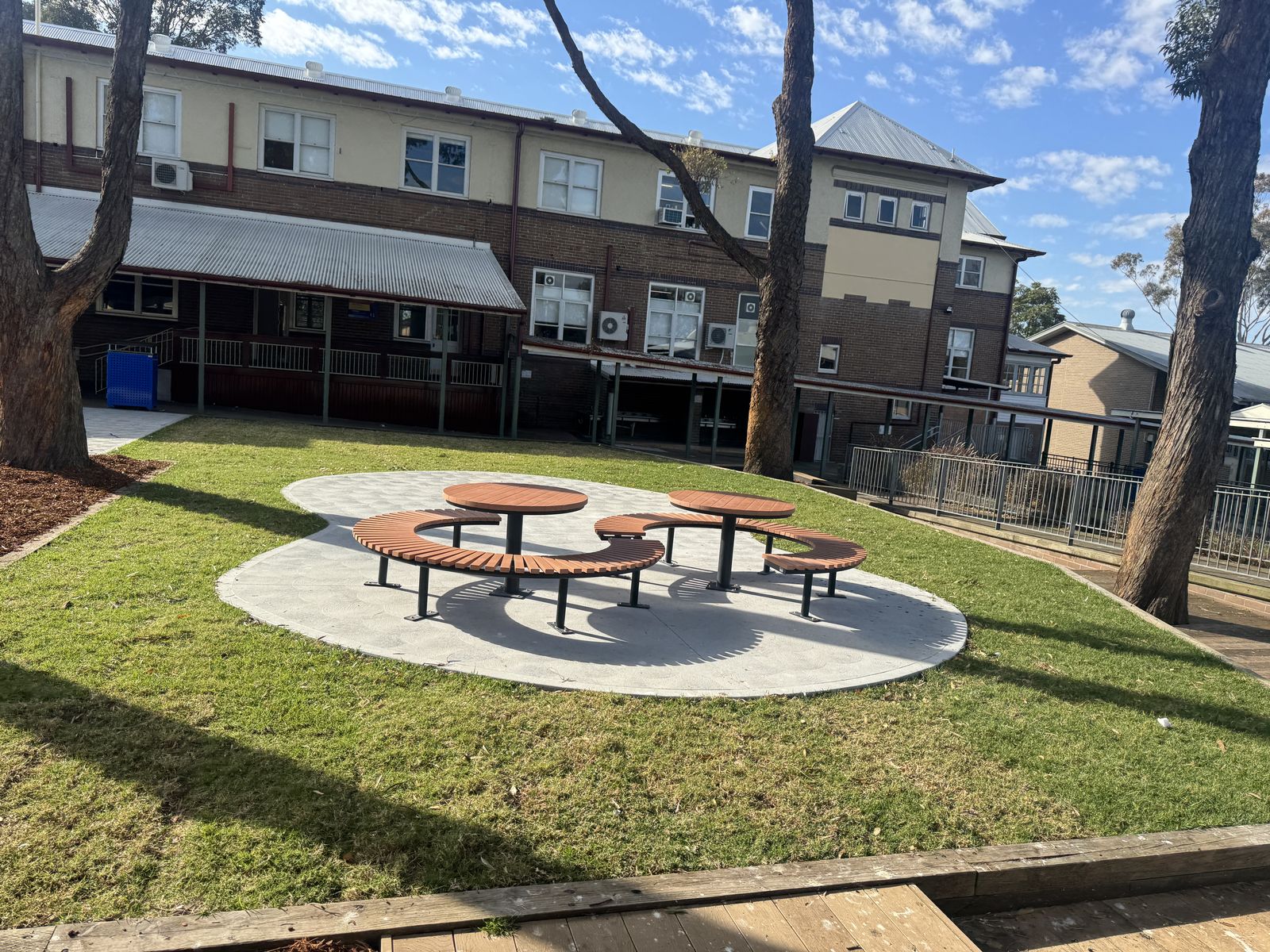 Upgraded courtyard with curved timber bench seating and tables on a round concrete pad, surrounded by new turf and garden mulch