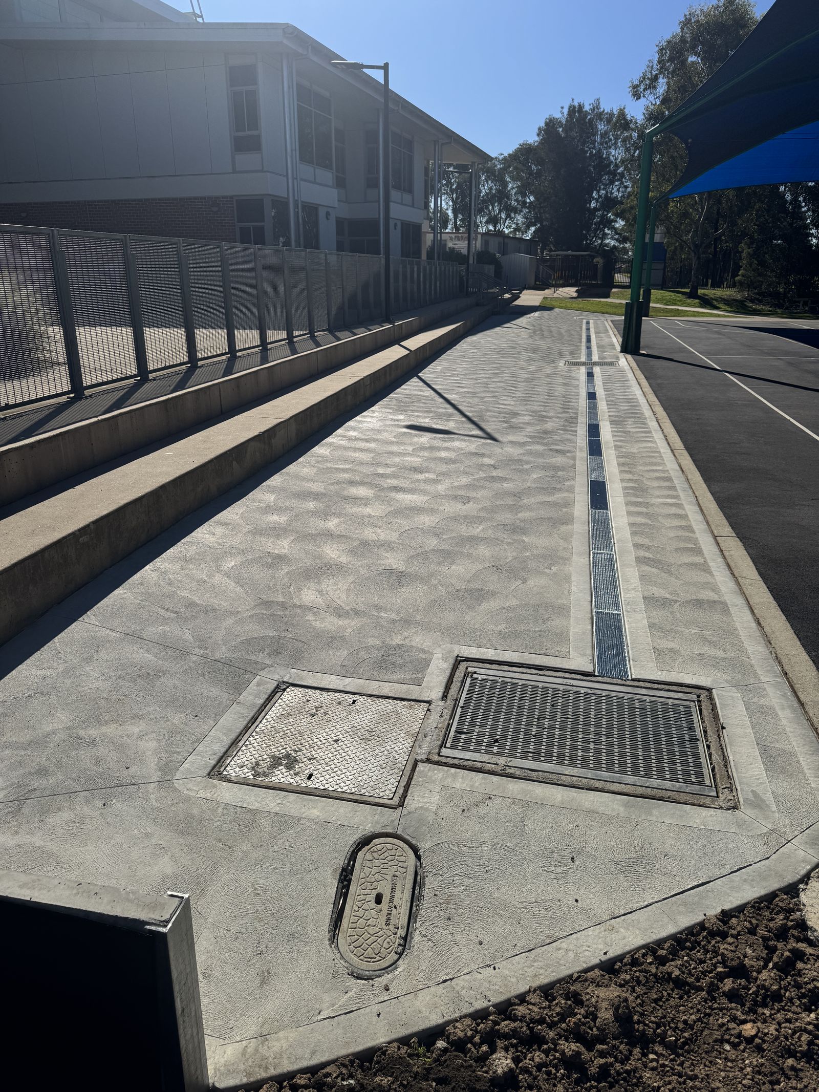 New stamped concrete path with a long grated drain and pit covers beside metal fencing